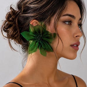 May include: A large, dark green fabric flower ear cuff featuring layered, translucent petals and a central cluster of small, matching green beads. The accessory is designed to sit prominently over the ear, creating a bold, floral-inspired statement piece.