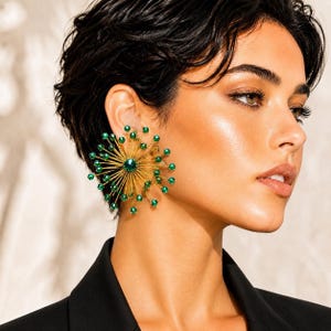 May include: A gold-toned, starburst-shaped earring with a central green gem and radiating green beads. The earring is worn on the ear, showcasing its intricate design and vibrant color against a dark background. The jewelry piece adds a touch of glamour.