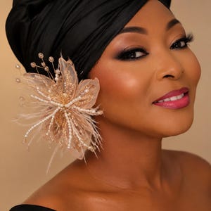 May include: A decorative hair accessory featuring a light peach-colored fabric flower with delicate feathers, small white pearls, and thin gold wire stems with clear beads, attached to a smooth black satin headwrap worn on the head.