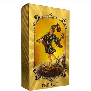 May include: A gold-colored tarot card box with a textured finish. The front features an illustration of a figure with a staff and a dog, set against a yellow background. The text "THE FOOL" is at the bottom.