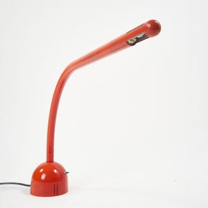 May include: A red desk lamp with a curved arm and a rounded head. The lamp has a circular base and a small light fixture at the end of the arm. The lamp is made of metal and has a glossy finish. The lamp is plugged into a black power cord.