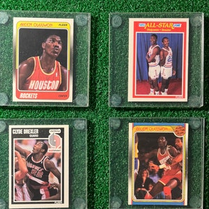 May include: Four vintage basketball trading cards featuring Akeem Olajuwon and Clyde Drexler. The cards are in protective cases and show players in action, with team logos and the text "Houston Rockets", "Blazers", and "1989 All-Star Game".