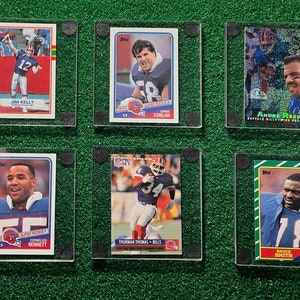 Może przedstawiać: Sześć kolekcjonerskich kart do gry w futbol amerykański z zawodnikami Buffalo Bills, prezentowanych w przezroczystych etui ochronnych. Karty przedstawiają graczy takich jak Jim Kelly, Thurman Thomas i Bruce Smith. Karty są na zielonym tle.