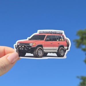 May include: A sticker of a red and white SUV with a roof rack and spare tire. The vehicle has black accents and large off-road tires. The sticker is held against a bright blue sky.