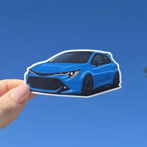 May include: A blue car sticker depicting a modern hatchback in a stylized illustration. The sticker has a white border and is held against a clear blue sky. The car has black wheels and a black front grill.