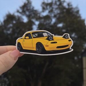 May include: A yellow sports car sticker with black wheels and pop-up headlights. The car is a stylized illustration with a white border, perfect for car enthusiasts. The sticker is held up against a blurred background of trees.