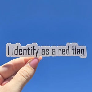 以下が含まれることがあります： 鮮やかな青い空を背景に、黒い文字で「I identify as a red flag」と書かれた白いステッカー。ステッカーは長方形で、角が丸くなっています。