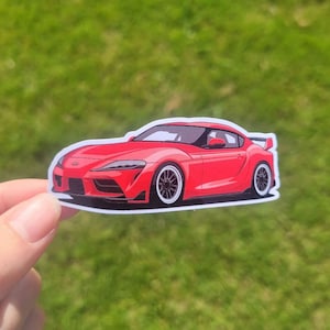 Puede incluir: Una pegatina de coche roja con una ilustración detallada de un coche deportivo moderno con un parachoques delantero negro y faldones laterales negros. El coche tiene ruedas negras con llantas plateadas y un alerón trasero negro.