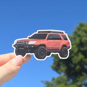May include: A sticker featuring a red SUV with black wheels and a roof rack. The vehicle is depicted in a detailed illustration, set against a bright blue sky. The sticker is held by a hand.