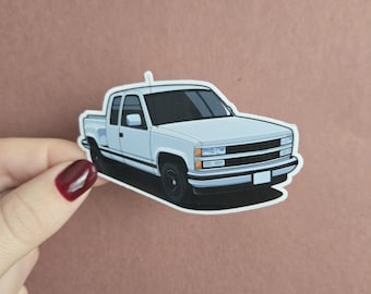 Puede incluir: Una pegatina azul claro de una camioneta con ruedas y molduras negras. La camioneta tiene una cabina y una caja blancas, con una sombra negra debajo. La pegatina está sujeta por una mano con esmalte de uñas granate.