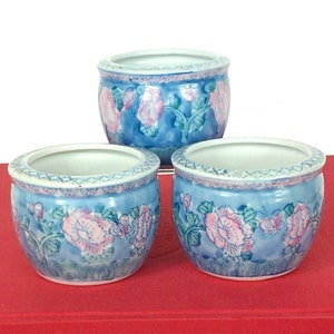 May include: Three small, round ceramic pots with a blue background and pink floral design. The pots are arranged on top of a red book. The pots have a white interior and a decorative rim.