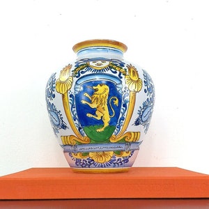 May include: A white ceramic vase with a blue and yellow floral design and a gold lion crest. The vase is sitting on a red surface.