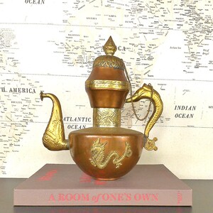 May include: A copper and gold teapot with a dragon design and a spout shaped like a dragon's head. The teapot is sitting on a book titled "A Room of One's Own".