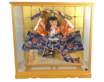 Op de afbeelding: Een Japanse pop in een houten vitrine. De pop draagt een kleurrijke traditionele outfit met een zwaard. De achtergrond is goudkleurig met een wit en rood touw. De vitrine heeft een glazen voorkant.