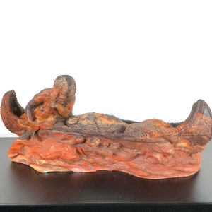 May include: A hand-carved wooden sculpture of a Native American in a canoe. The sculpture is painted in shades of red, brown, and black.