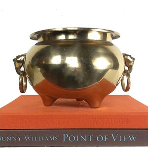 May include: A gold brass planter with lion head handles and three legs. The planter is sitting on top of two books, one orange and one brown. The brown book has the text "Bunny Williams' Point of View" on the spine.