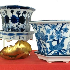 May include: Three ceramic planters with blue and white floral designs. One planter features a lotus design, while another showcases birds and cherry blossoms. A gold-colored sculpture and two books are also in the image.