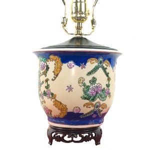May include: A vintage ceramic table lamp with a floral design. The lamp features a cream-colored body with blue, pink, and gold floral patterns. It has a brass lamp holder and a dark wooden base. The lamp is approximately 20 inches tall.