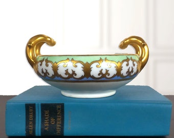May include: A white porcelain bowl with gold accents and a green and blue design. The bowl has two gold handles and is sitting on a teal blue book.