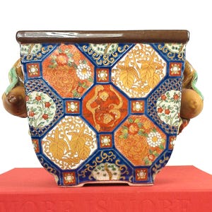 May include: A square ceramic planter with a detailed, colorful design. The planter features a brown rim, blue borders, and orange, yellow, and white floral and geometric patterns. Two fruit-shaped handles are on the sides. The planter sits on a red book.