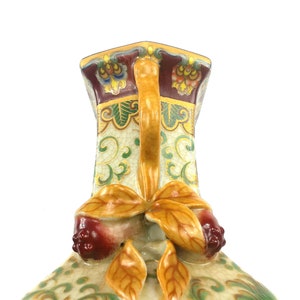 May include: A ceramic vase with a square base and a decorative handle. The vase is decorated with a floral pattern in shades of green, yellow, and red.