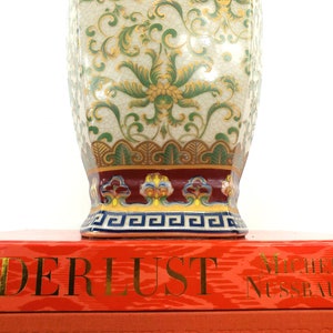 May include: A decorative porcelain vase with a white background and green and gold floral pattern. The vase has a red and gold band around the top and a blue and gold band around the bottom. The vase is sitting on top of a red book with the title "Derlust" and the author "Michel Nussbaumer".