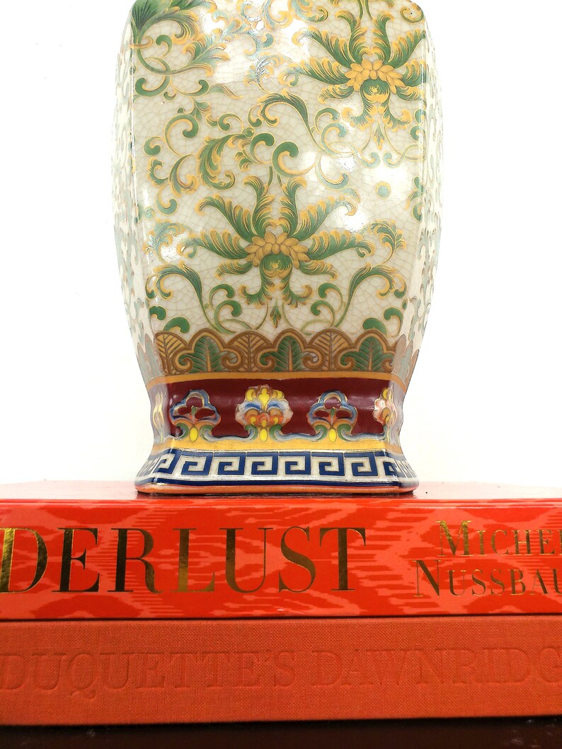 May include: A decorative porcelain vase with a white background and green and gold floral pattern. The vase has a red and gold band around the top and a blue and gold band around the bottom. The vase is sitting on top of a red book with the title "Derlust" and the author "Michel Nussbaumer".