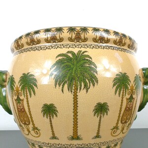 May include: A large, cream-colored ceramic planter with green and gold palm tree designs. The planter has two green, sculpted handles in the shape of a face.