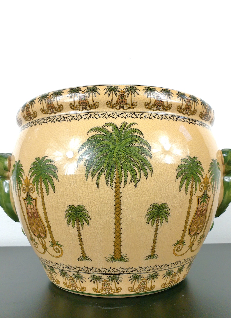 May include: A large, cream-colored ceramic planter with green and gold palm tree designs. The planter has two green, sculpted handles in the shape of a face.