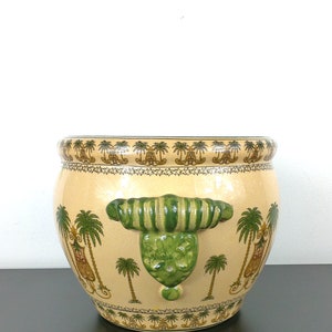 May include: A large, cream-colored ceramic planter with green and gold palm tree designs. The planter has two green, striped handles on the sides.
