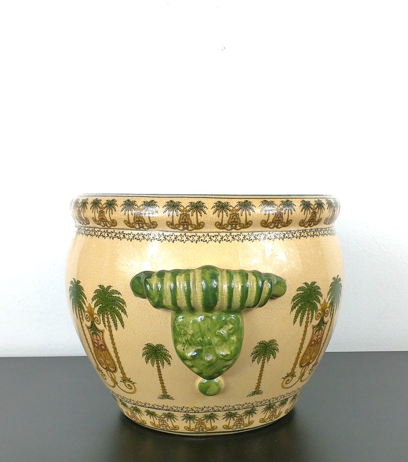 May include: A large, cream-colored ceramic planter with green and gold palm tree designs. The planter has two green, striped handles on the sides.