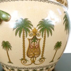 May include: A large, cream-colored ceramic planter with a green handle and a detailed design of palm trees and ornate scrollwork. The planter has a crackled glaze and a border of small stars.