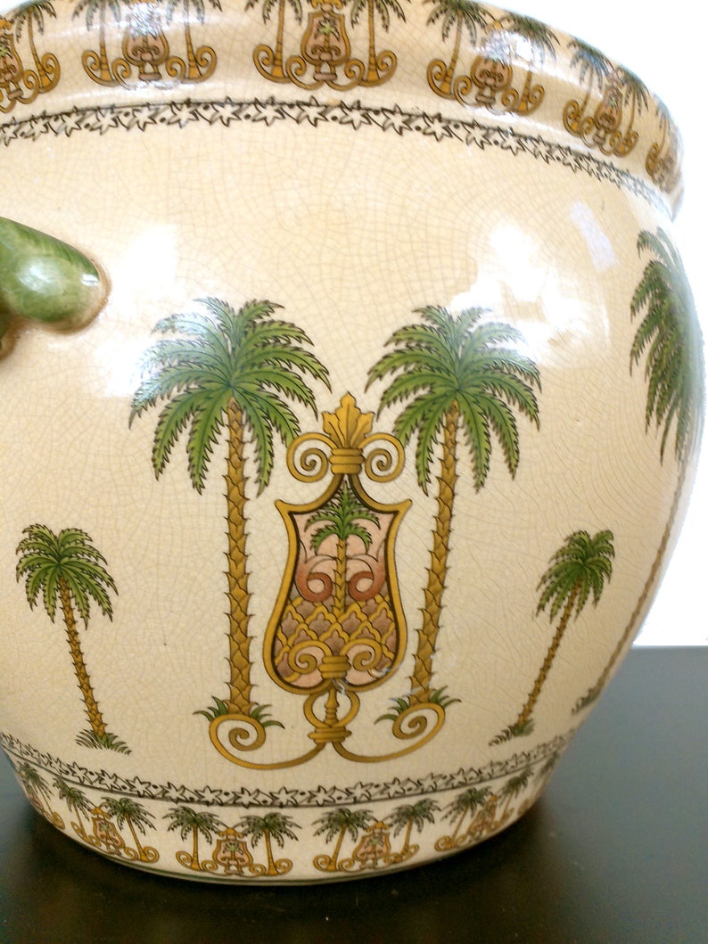 May include: A large, cream-colored ceramic planter with a green handle and a detailed design of palm trees and ornate scrollwork. The planter has a crackled glaze and a border of small stars.