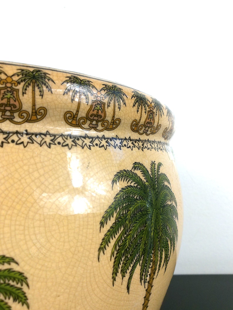 May include: A large, cream-colored ceramic planter with a crackled glaze. The planter is decorated with a repeating pattern of palm trees and a border of stars. The planter is perfect for adding a touch of tropical flair to any home.