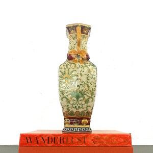 May include: A decorative ceramic vase with a green, yellow, and red floral pattern. The vase has a square base and a round body. It is sitting on top of two orange books with the word "WANDERLUST" printed on the top book.