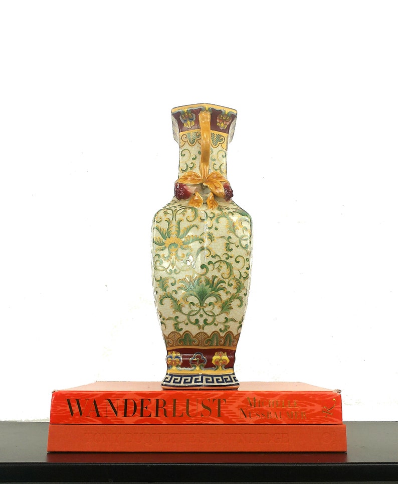 May include: A decorative ceramic vase with a green, yellow, and red floral pattern. The vase has a square base and a round body. It is sitting on top of two orange books with the word "WANDERLUST" printed on the top book.