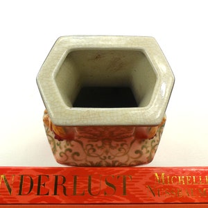 May include: A hexagonal ceramic vase with a white glaze and a floral pattern in shades of orange, green, and gold. The vase has a wide opening and a slightly flared lip.