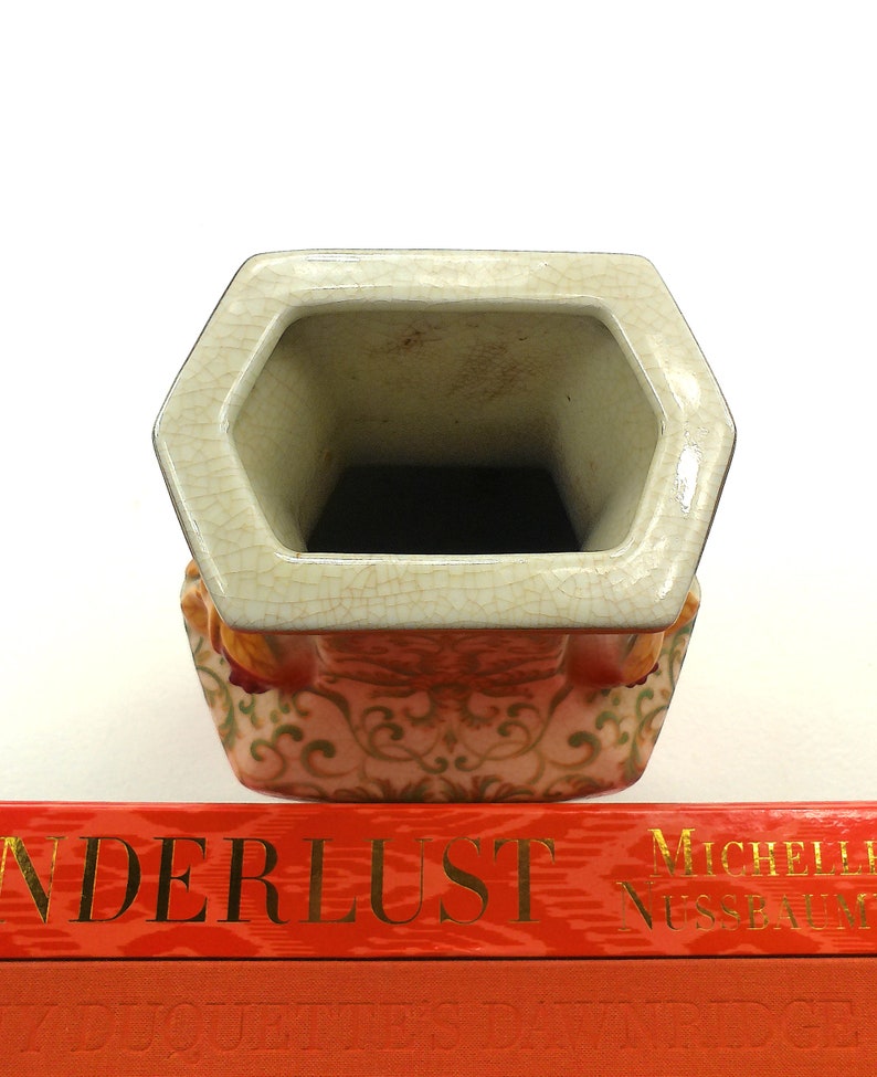 May include: A hexagonal ceramic vase with a white glaze and a floral pattern in shades of orange, green, and gold. The vase has a wide opening and a slightly flared lip.