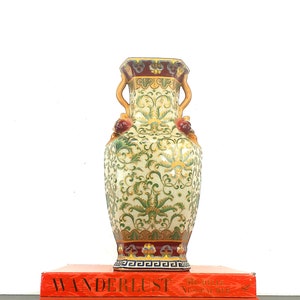 May include: A decorative ceramic vase with a green and gold floral pattern, red accents, and a gold rim. The vase has two handles and is sitting on top of a red book with the word "WANDERLUST" printed on the cover.