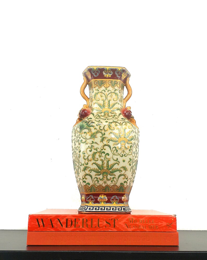 May include: A decorative ceramic vase with a green and gold floral pattern, red accents, and a gold rim. The vase has two handles and is sitting on top of a red book with the word "WANDERLUST" printed on the cover.
