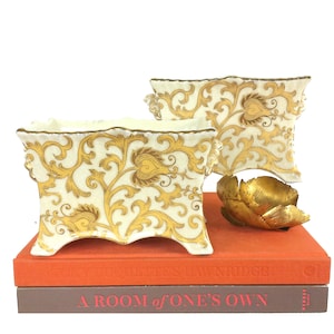 May include: Two cream-colored rectangular planters with gold floral designs and a gold-colored metal flower sculpture rest on a stack of books. The books are orange and brown, with the title "A Room of One's Own" visible.
