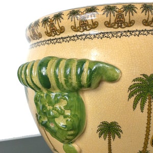 May include: A large, cream-colored ceramic planter with green and brown palm tree designs. The planter has a unique, green, three-dimensional, ribbed handle on one side.