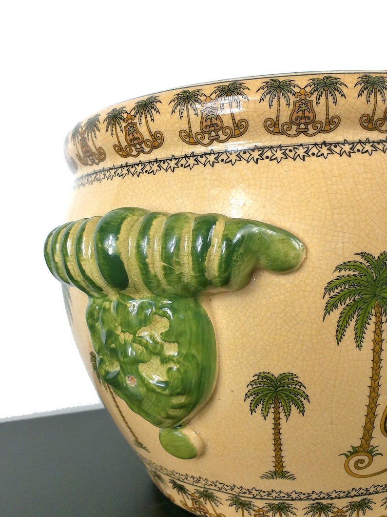 May include: A large, cream-colored ceramic planter with green and brown palm tree designs. The planter has a unique, green, three-dimensional, ribbed handle on one side.