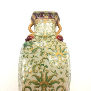 May include: A white porcelain vase with green and gold floral designs and a brown and red rim. The vase has two handles shaped like stylized animal heads.