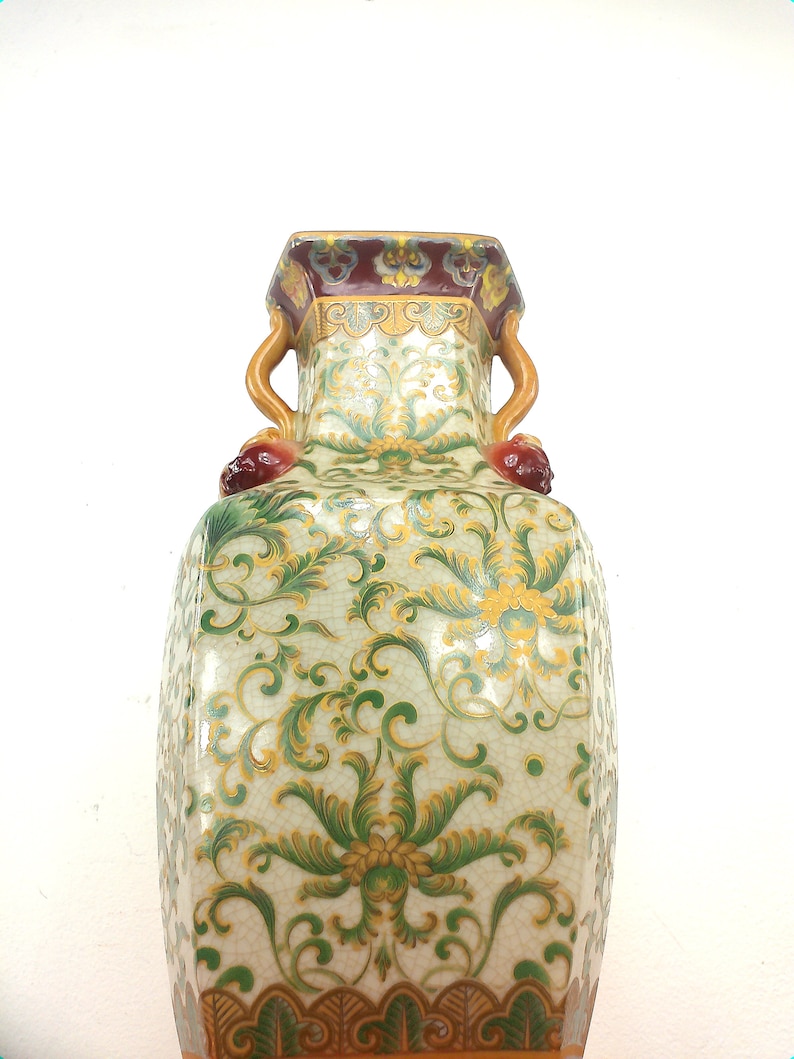 May include: A white porcelain vase with green and gold floral designs and a brown and red rim. The vase has two handles shaped like stylized animal heads.