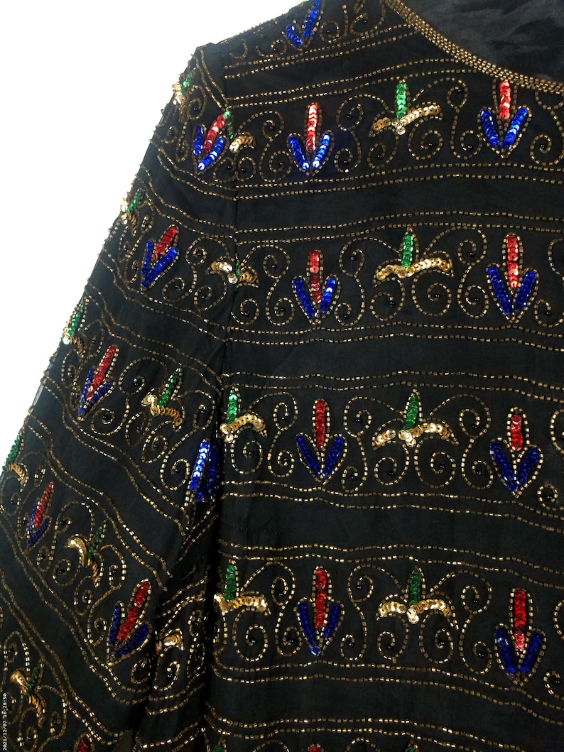 Pu&ograve; includere: Una giacca nera di paillettes con un motivo ripetuto di paillettes dorate, blu, rosse e verdi. Le paillettes sono disposte in un design floreale.