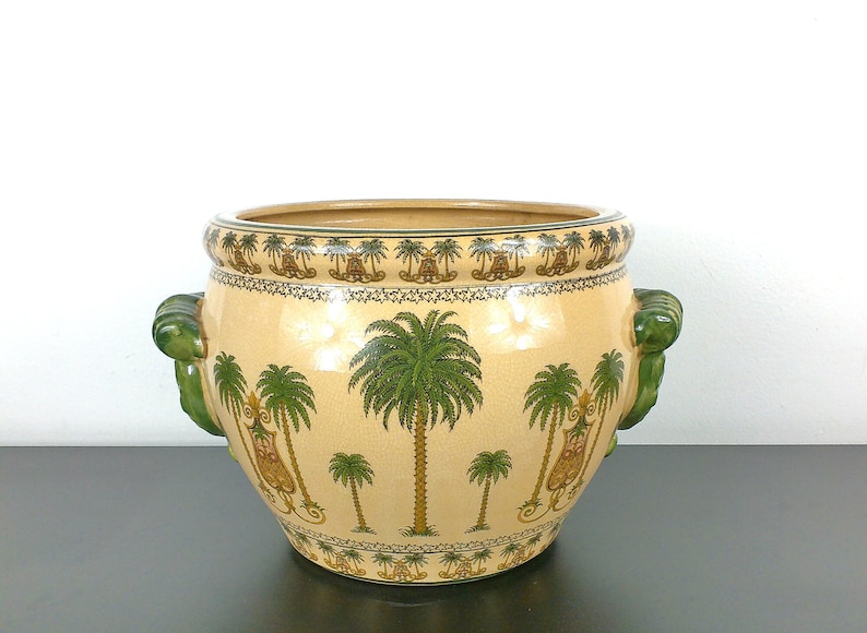 May include: A ceramic planter with green handles and a beige background. The planter is decorated with a repeating pattern of palm trees and small figures.