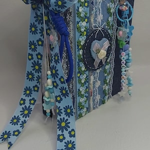 May include: A handmade, blue fabric-covered journal with floral ribbon and lace accents. The journal features a denim heart applique and beaded charms. A large bow with daisy pattern ribbon adorns the top. The overall design is a mix of textures and embellishments.