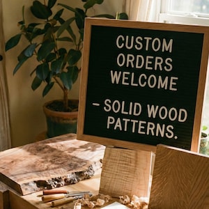 Könnte beinhalten: Ein schwarzes Filz-Buchstabenbrett mit einem hellen Eichenholzrahmen, das den weißen Text CUSTOM ORDERS WELCOME - SOLID WOOD PATTERNS anzeigt. Das Brett steht auf einem Holzständer neben einer Holzplatte mit Naturkante und zwei Schnitzwerkzeugen.