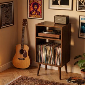 May include: A dark wood record player stand with four tapered legs, featuring an open top shelf holding a turntable and a Marshall speaker, and a lower shelf filled with a collection of vinyl records arranged vertically in a row.